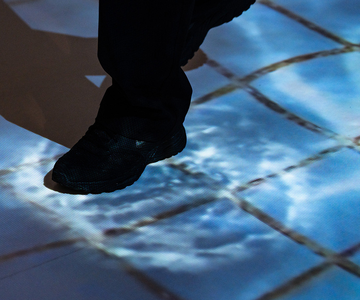 床には、水が張ったタイルのような映像が映し出されている。黒い靴を履いた人の足元は、水の波紋のような映像が映し出されている。