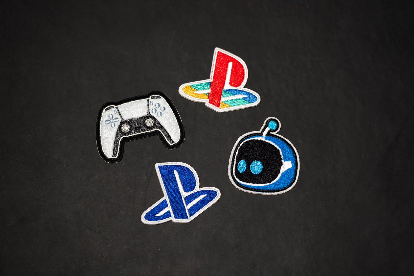 Photo of the original patch stickers. The top left features a DualSense wireless controller, the bottom left has a simple design in solid blue, the bottom right showcases a motif of 'Astro Bot's' face, and the top right recreates the colored logo of the original PlayStation family mark in red, yellow, green, and blue.