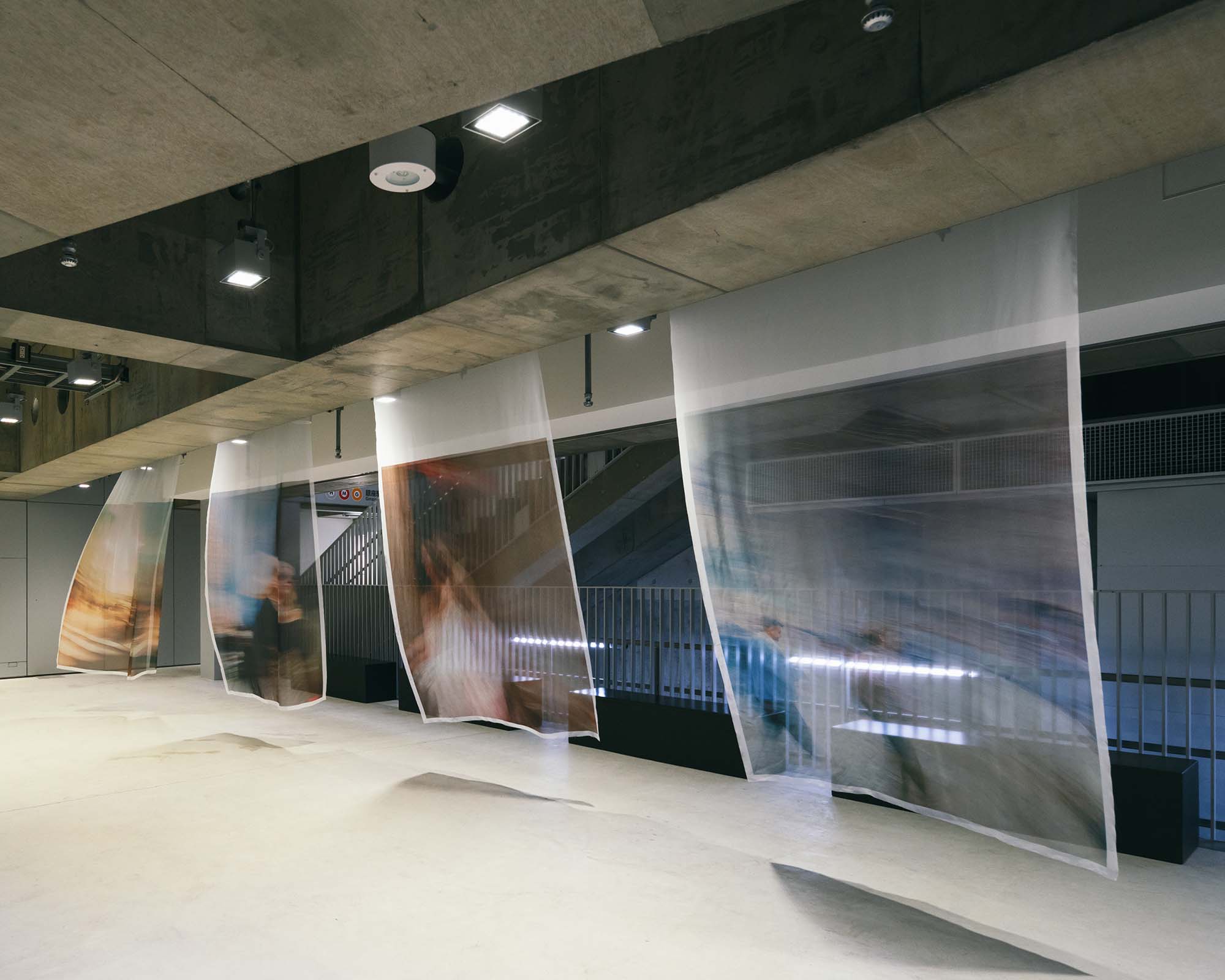 At Ginza Sony Park B1, multiple translucent panels hang in sequence, blending blurred figures with the concrete architecture.