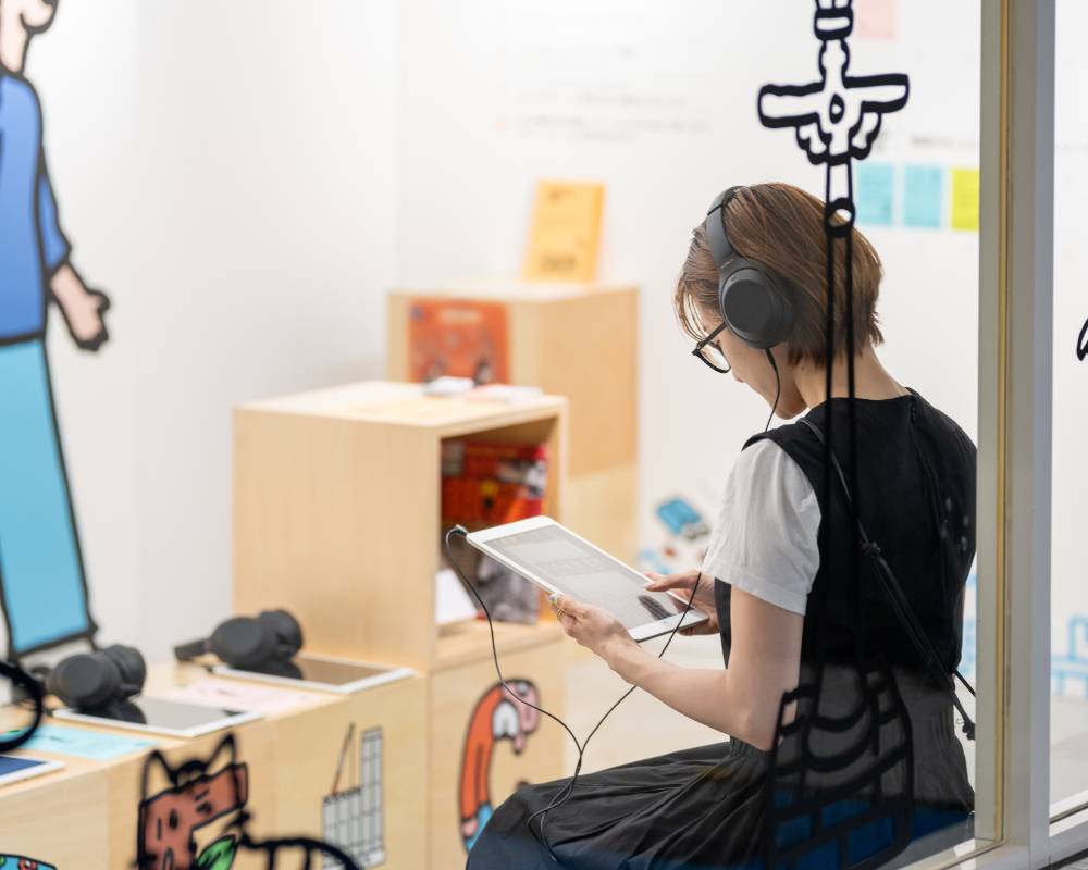 体験の様子。来場者がベンチに着席し、ヘッドフォンをつけてタブレットを眺めている。