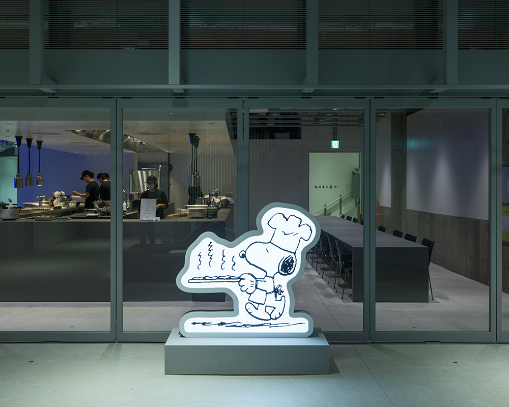 A glowing sign of Snoopy dressed as a chef in front of a restaurant kitchen visible through glass walls.