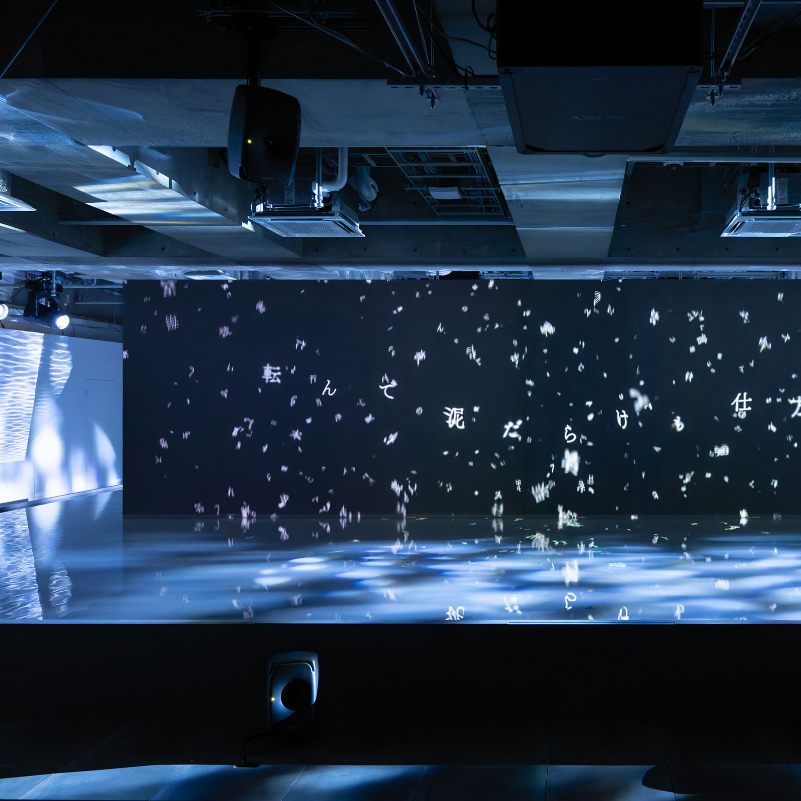 展示風景。黒い壁面にプロジェクションされるさまざまな言葉と、その手前のある水面と光の反射によるインスタレーション。