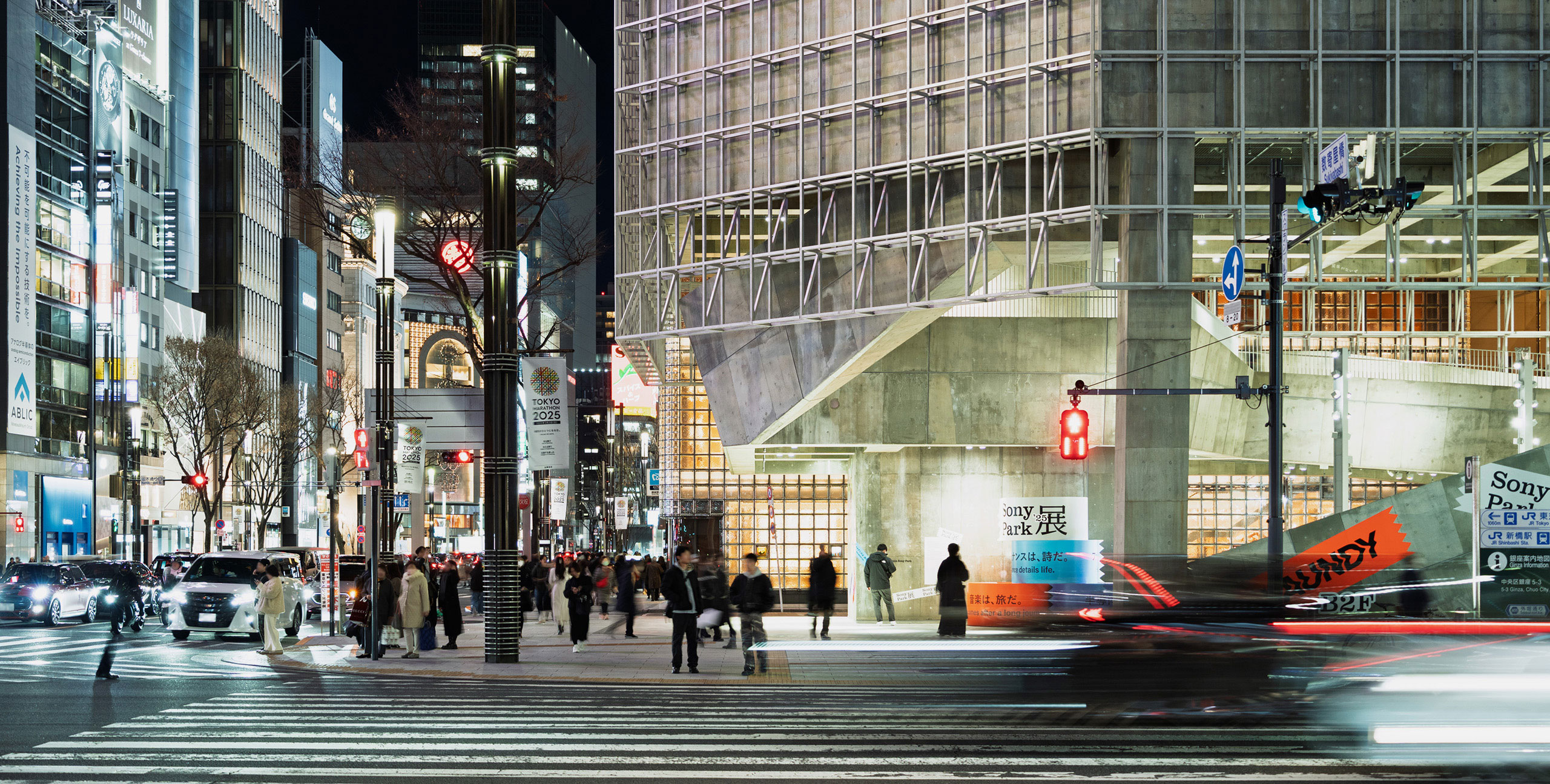 A nighttime cityscape at a street corner where many people and cars are waiting at a traffic light.