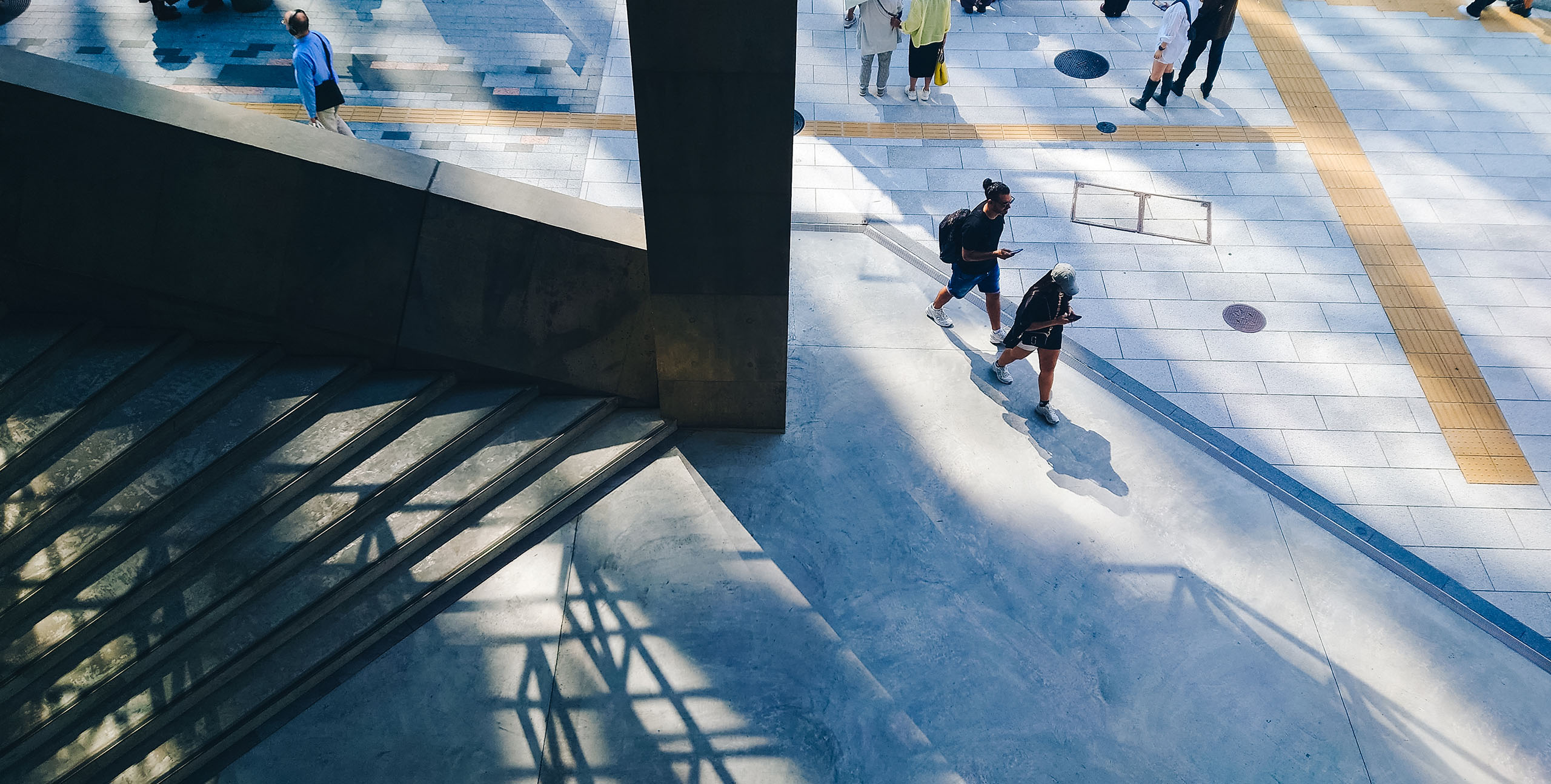 A stairwell at Ginza Sony Park. Pedestrians walk on the pavement above as sunlight and shadows intersect across the concrete surfaces.
