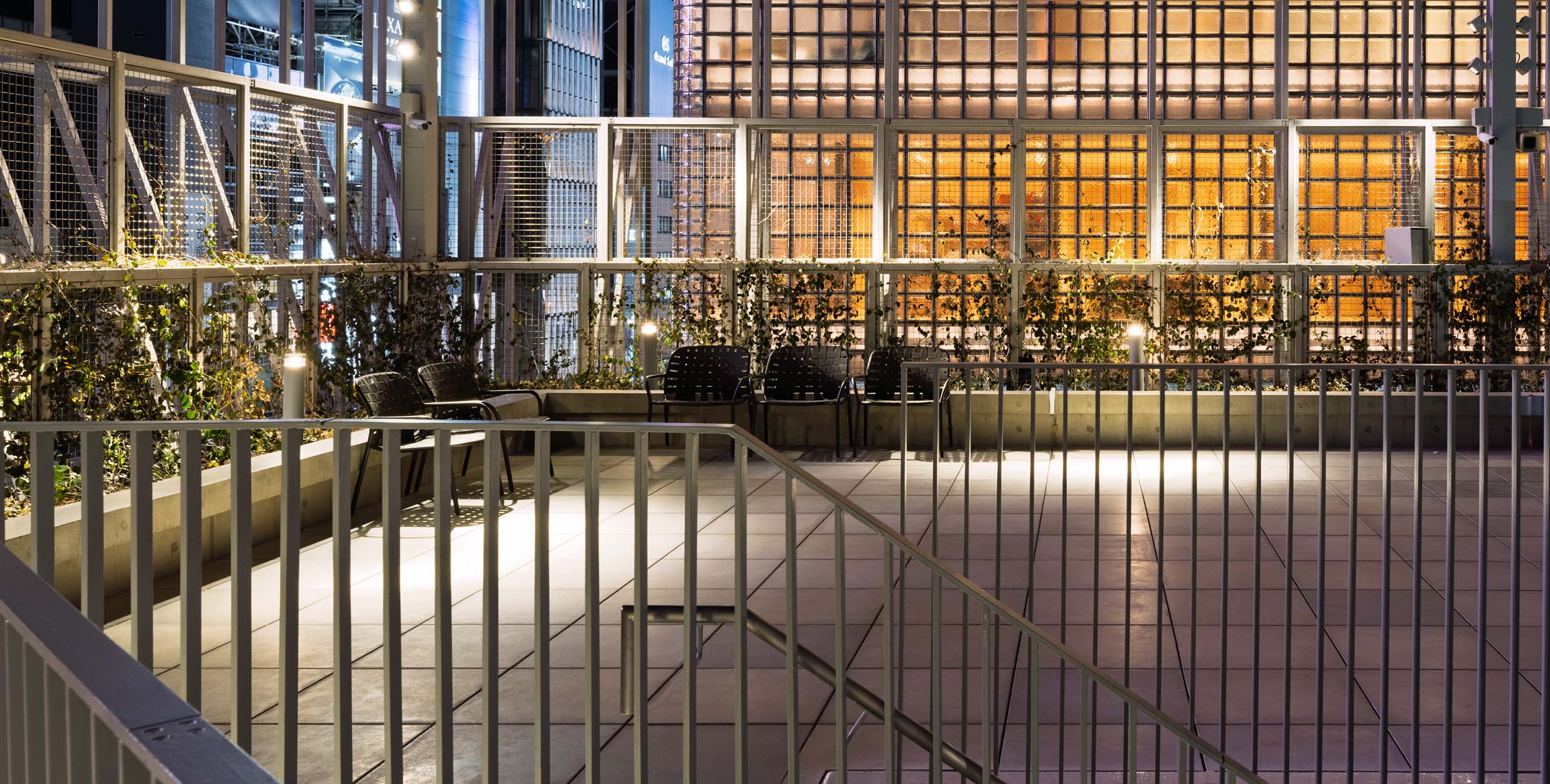 Night view of the rooftop terrace at Ginza Sony Park. Warm light filters through the glass-block walls, illuminating a tranquil space with greenery and arranged chairs.