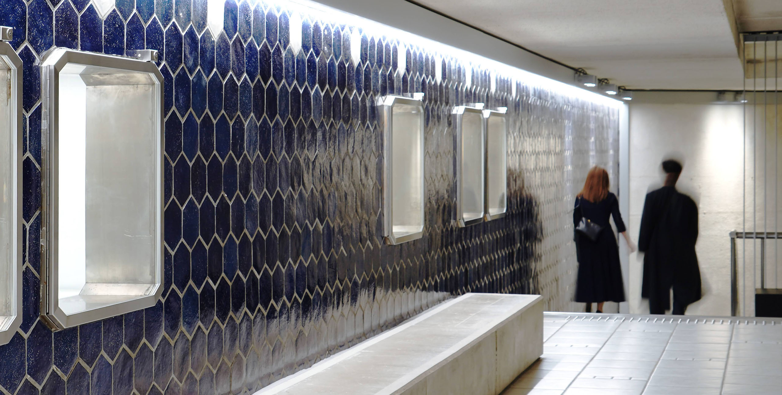 A blue-tiled corridor leading underground at Ginza Sony Park. The hexagonal tiles reflect the light, while a couple walks ahead in the softly illuminated passage.