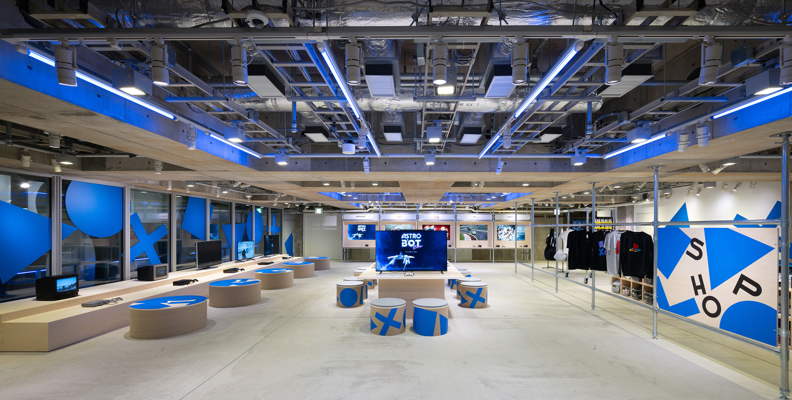 Ginza Sony ParkのB2に、▲●✕■をモチーフにした青いグラフィックが壁や椅子に展開されている。中央にはゲーム体験用の大型モニターが置かれ、右側にはアパレルやグッズが並ぶショップが続く。
