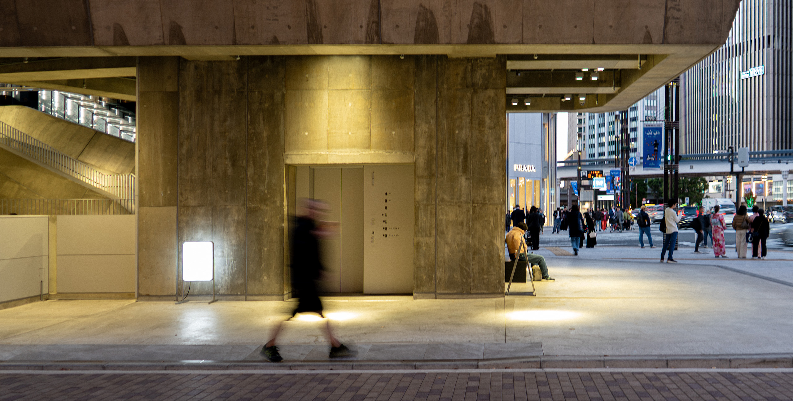 夕方のGinza Sony Park1Fの入口付近。打ち放しコンクリートの柱が立ち並ぶ下、歩行者が行き交い、奥には明るい商業ビルと交差点の人だかりが見える。