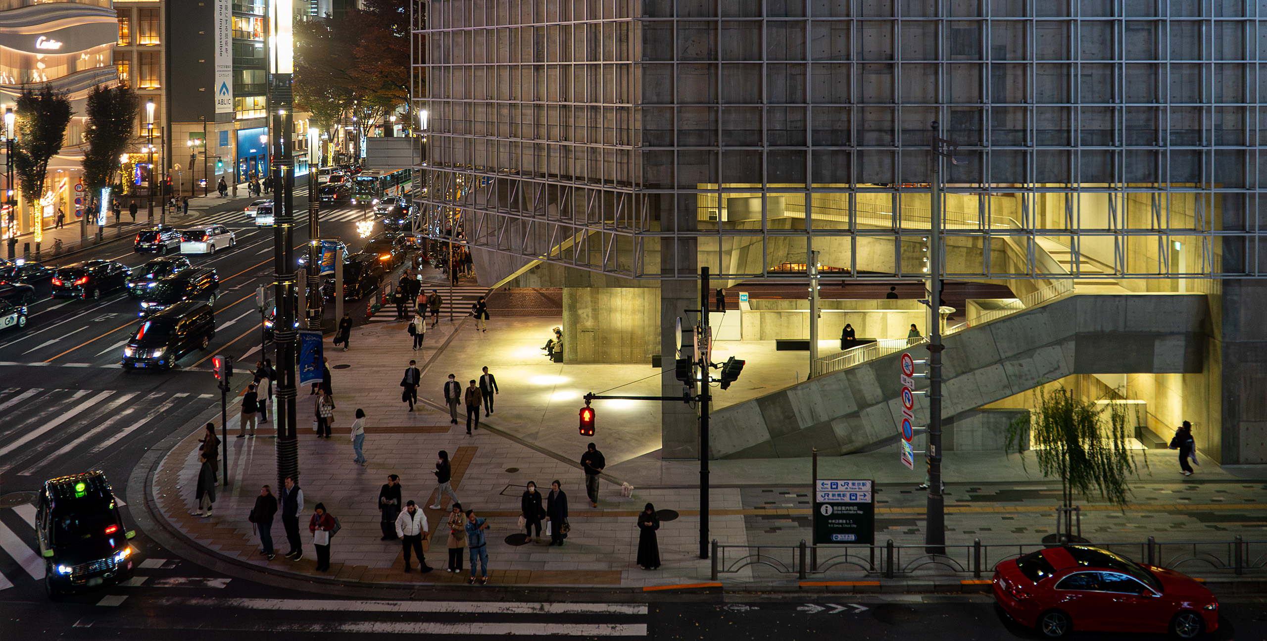 夜の銀座四丁目交差点を見下ろす構図。車のライトが街路を流れ、Ginza Sony Parkの低層部では明るく照らされた階段空間に人々が佇む。