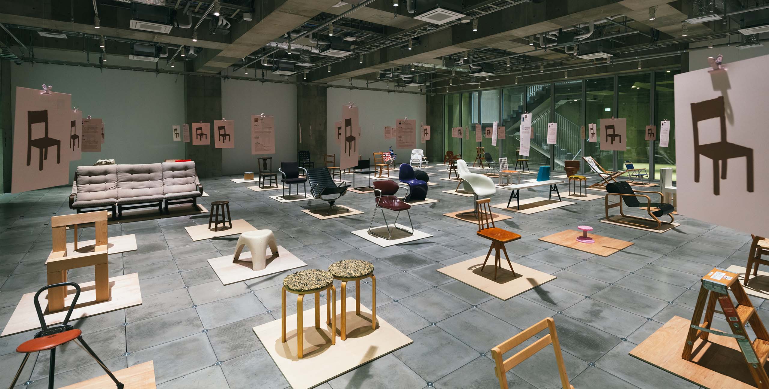 Ginza Sony Parkの展示フロアに、多彩な椅子が等間隔に並ぶ風景。吊り下げられた図版とコンクリート空間が、デザインの対話を生み出している。